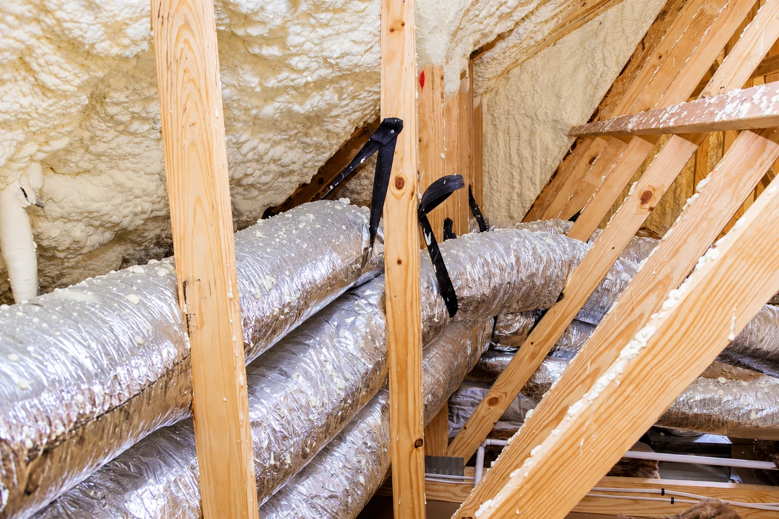 Foam insulation coats the roof framing around shiny flexible ducts in an attic.