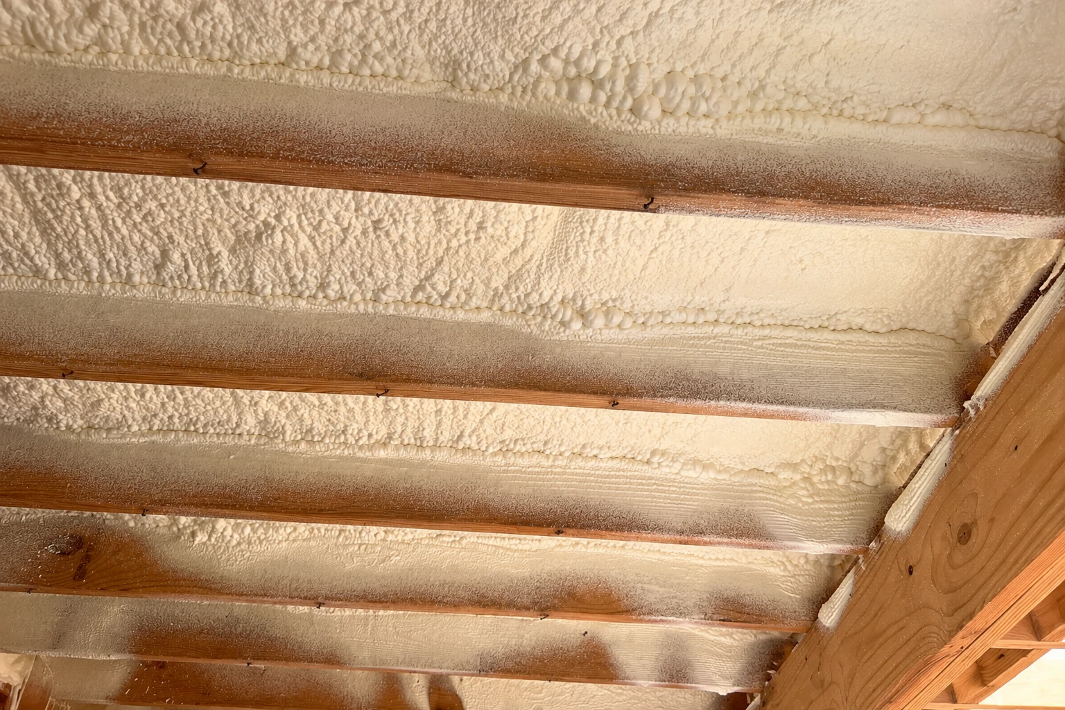 Close view of white spray foam filling the spaces between wooden ceiling joists.