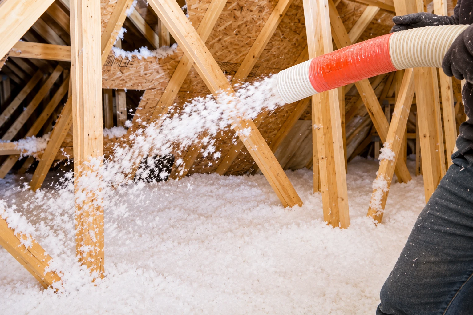 A worker blows loose white insulation through a hose across an attic floor between exposed wooden trusses.