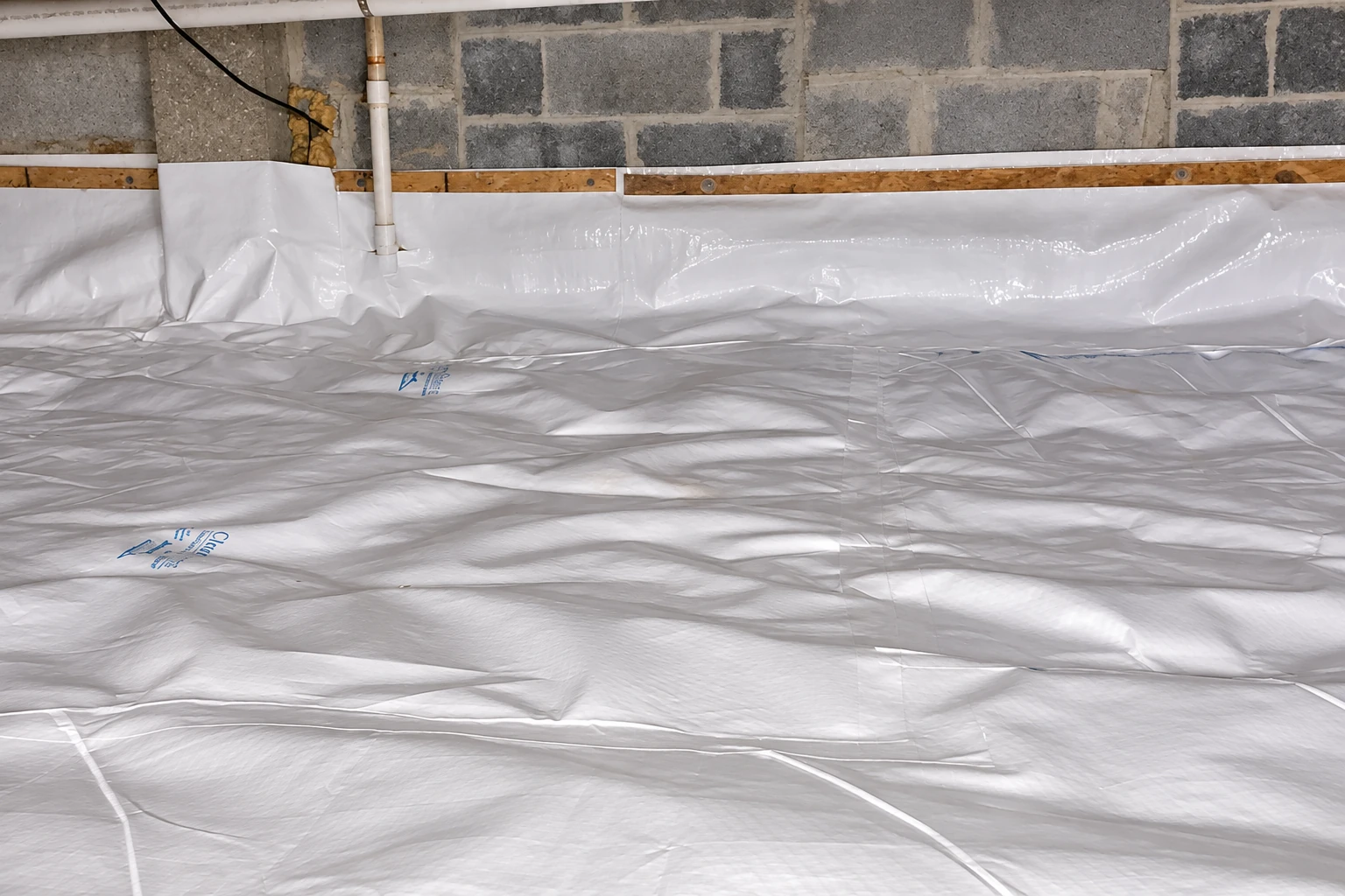 Crawl space floor covered with overlapping white plastic vapor barrier beneath a block wall and exposed pipes.