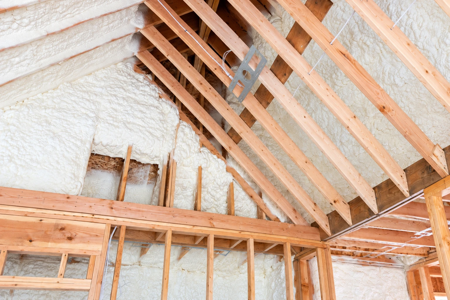 White spray foam insulation coats the underside of a roof and walls around exposed wooden attic framing.