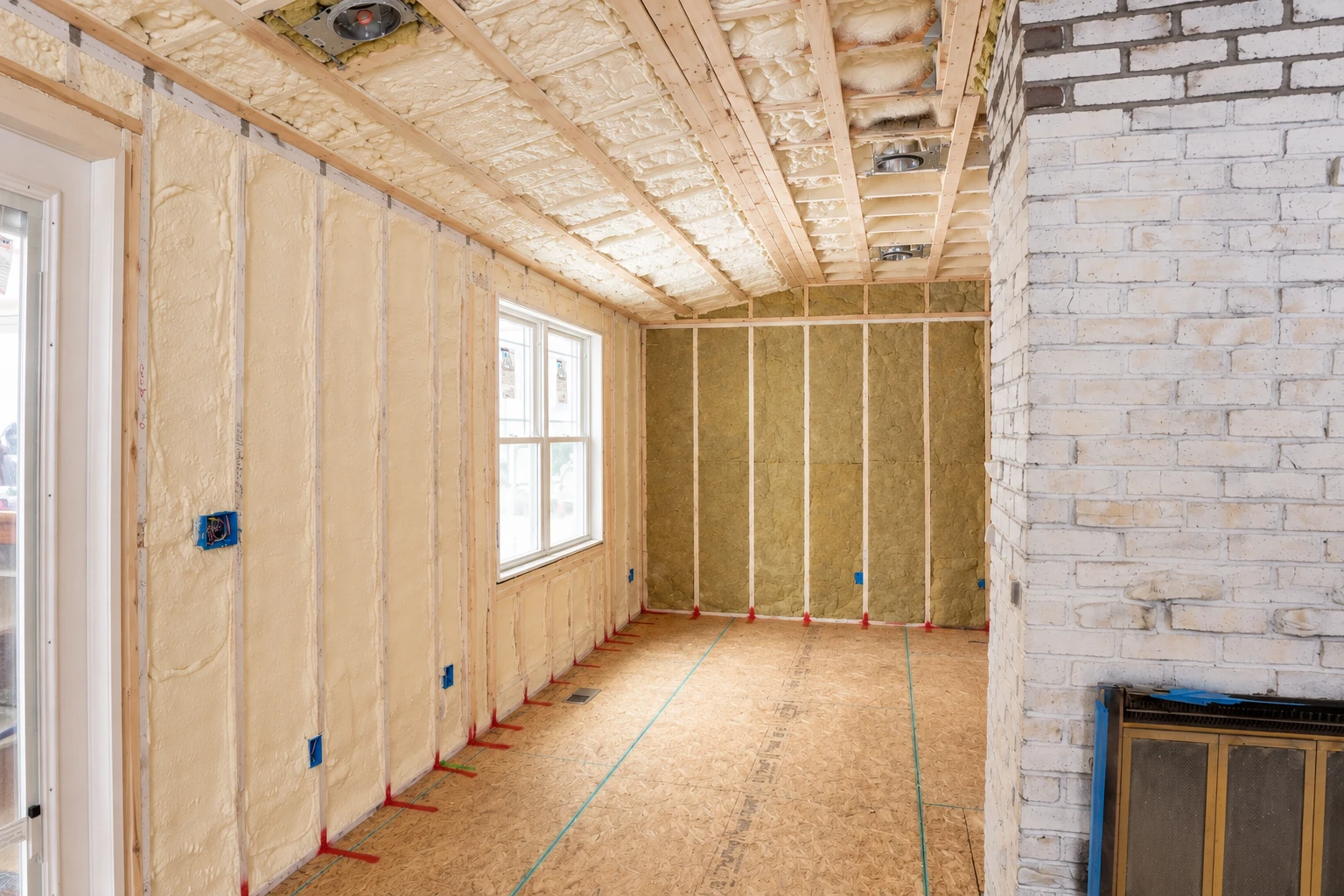 Spray foam fills the wall and ceiling cavities of a framed room beside a white brick fireplace.